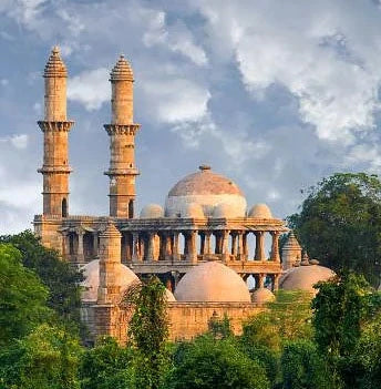 An old domed stone mosque with two tall minarets, rising above green trees.